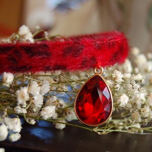 May include: A red faux fur collar with a dangling red gemstone pendant. The collar is trimmed with black spots and the pendant is attached to a gold chain.