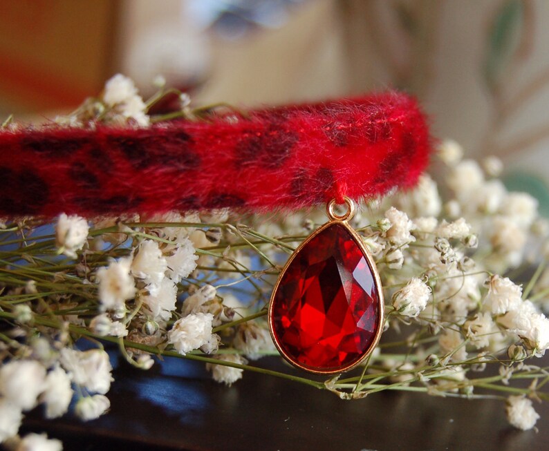 May include: A red faux fur collar with a dangling red gemstone pendant. The collar is trimmed with black spots and the pendant is attached to a gold chain.