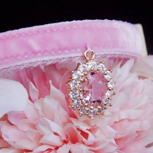 May include: A pink velvet cat collar with a gold pendant featuring a large oval-shaped pink gemstone surrounded by clear rhinestones.