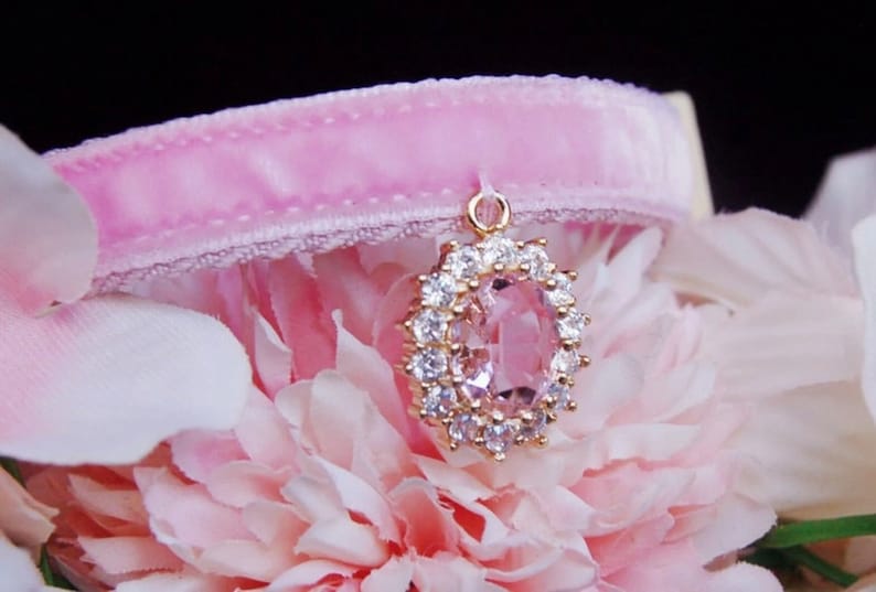 May include: A pink velvet cat collar with a gold pendant featuring a large oval-shaped pink gemstone surrounded by clear rhinestones.