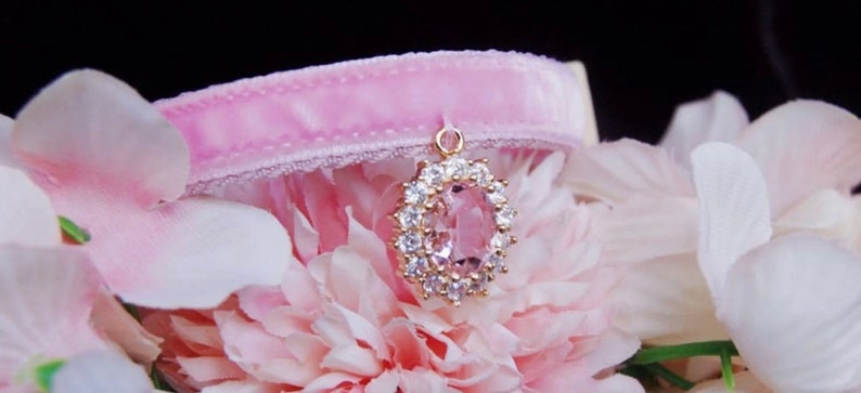 May include: A pink velvet choker with a gold pendant featuring a large oval-shaped pink gemstone surrounded by smaller clear gemstones.