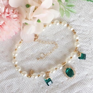 May include: A white pearl bracelet with a gold chain and three emerald green gemstone charms. The charms are different shapes, including a square, a rectangle, and an oval.