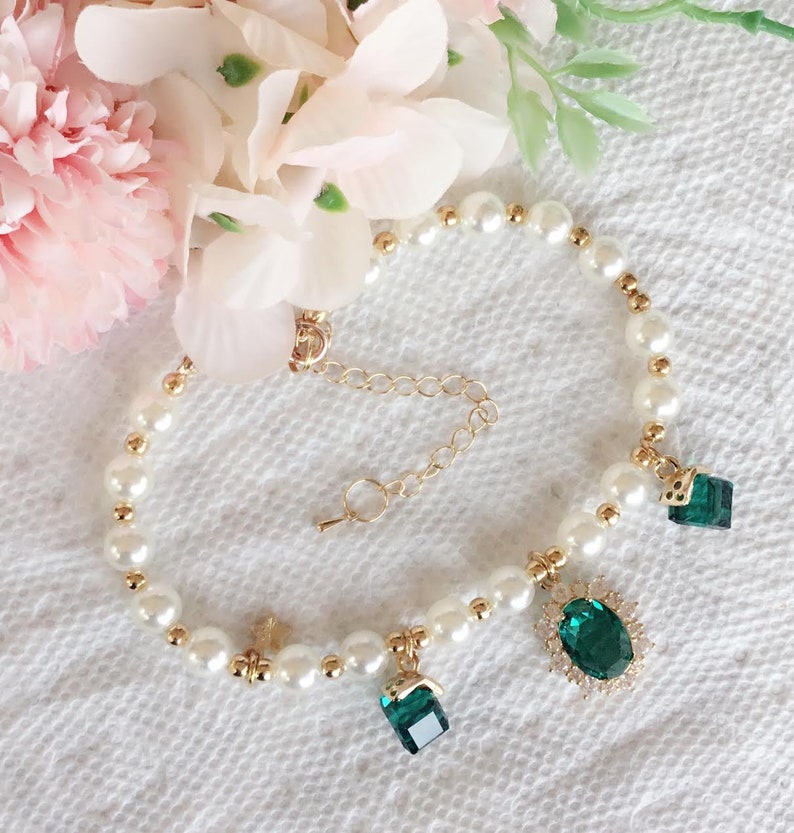 May include: A white pearl bracelet with a gold chain and three emerald green gemstone charms. The charms are different shapes, including a square, a rectangle, and an oval.