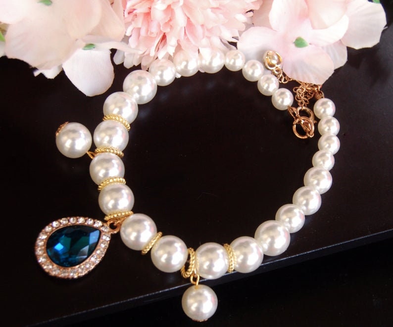 K&ouml;nnte beinhalten: Ein wei&szlig;es Perlenarmband mit einer goldenen Kette und zwei tropfenf&ouml;rmigen Anh&auml;ngern. Ein Anh&auml;nger ist ein gro&szlig;er blauer Edelstein, der von kleineren klaren Edelsteinen umgeben ist, der andere ist eine kleinere wei&szlig;e Perle.