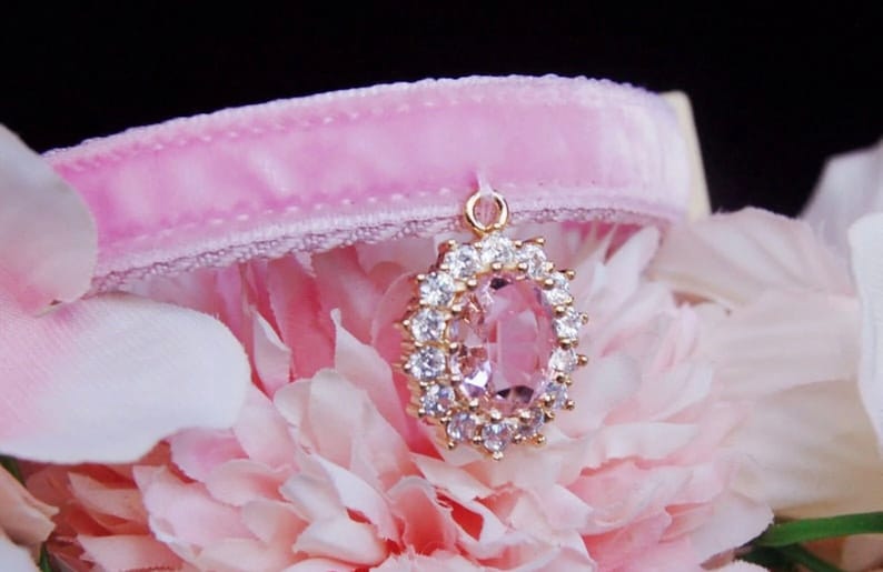 May include: A pink velvet cat collar with a gold pendant featuring a large pink crystal surrounded by smaller clear crystals.
