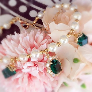 May include: A gold bracelet with white pearls and green gemstone charms. The bracelet is on a pink flower.