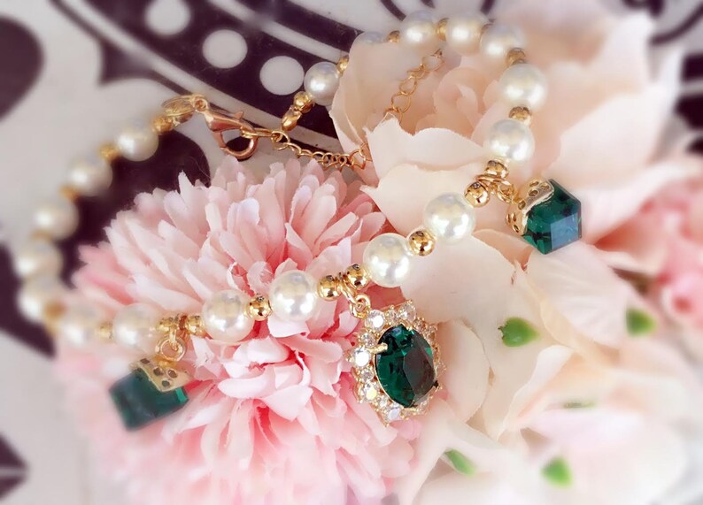 May include: A gold bracelet with white pearls and green gemstone charms. The bracelet is on a pink flower.
