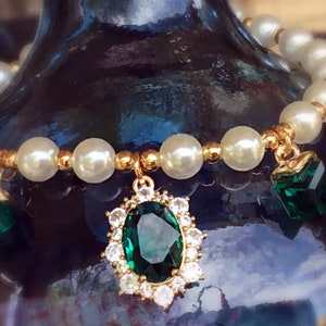 May include: A gold bracelet with white pearl beads and emerald green gemstone charms. The bracelet has a large emerald green gemstone pendant with a gold setting.