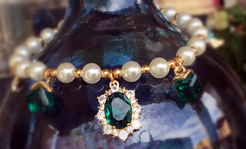 May include: A gold bracelet with white pearl beads and emerald green gemstone charms. The bracelet has a large emerald green gemstone pendant with a gold setting.