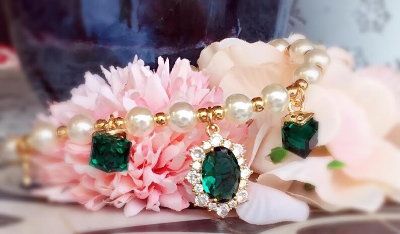 May include: A gold bracelet with white pearl beads and three green gemstone charms. The charms are a square, a round, and an oval shape.