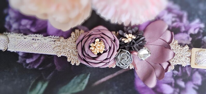 May include: A delicate white lace collar with a purple flower embellishment. The flower is made of fabric and has a layered design. The collar is adorned with small, sparkly accents.