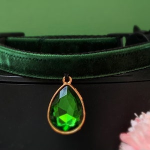 May include: A dark green velvet cat collar with a gold-tone pendant featuring a large, faceted, emerald green gemstone.