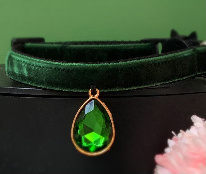 May include: A dark green velvet cat collar with a gold-tone pendant featuring a large, faceted, emerald green gemstone.