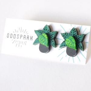 May include: A pair of plant-shaped earrings. Each earring has a dark grey pot with green leaves and a bright green leaf in the centre. The earrings are displayed on a white card with the text "ODD SPARK" in grey.