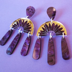 May include: A pair of statement earrings with a purple and gold color scheme. The earrings feature a teardrop-shaped top, a gold semi-circle with a dark purple floral design, and three dangling purple teardrops. The earrings are on a purple background.