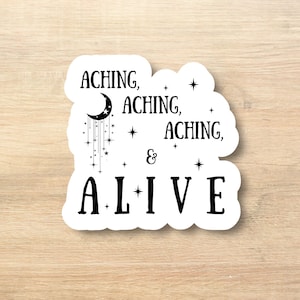 May include: A white sticker with black text and a crescent moon design. The text reads "ACHING, ACHING, ACHING, & ALIVE" with stars and a crescent moon with dangling stars. The sticker is on a light brown wooden surface.