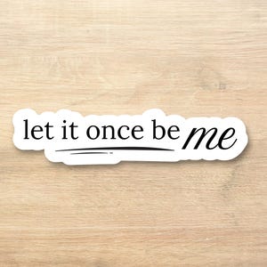 May include: A white sticker with the phrase "let it once be me" in a black, handwritten-style font. The sticker has a black underline beneath the words "let it once". The sticker is placed on a light brown wooden surface.