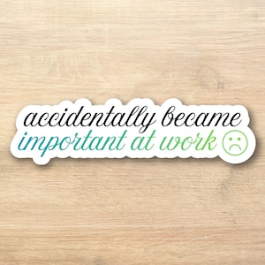 May include: A white sticker with the text "accidentally became important at work" in black and teal script. A green sad face is on the right side of the sticker. The sticker is on a light wood surface.