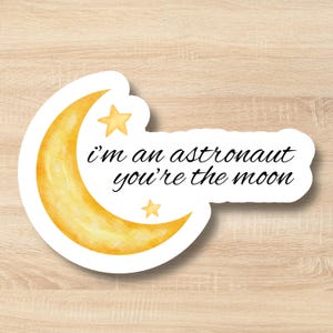 Puede incluir: Una pegatina troquelada con una luna creciente amarilla en acuarela y dos pequeñas estrellas amarillas. El texto negro impreso en la pegatina dice "i'm an astronaut you're the moon" en una fuente cursiva sobre un fondo blanco.