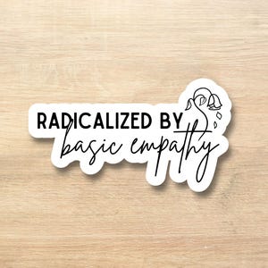 May include: A white sticker with the words "RADICALIZED BY basic empathy" in black script. The sticker has a floral design on the right side. The sticker is on a light brown wooden surface.