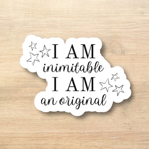 May include: A white sticker with black text and star illustrations. The text reads "I AM inimitable I AM an original". The sticker is on a light wood grain surface.
