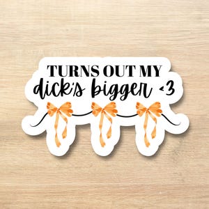 Puede incluir: Una pegatina blanca con el texto "TURNS OUT MY dick's bigger <3" en escritura negra. La pegatina presenta tres lazos de acuarela naranja conectados por una línea negra, creando un diseño juguetón.