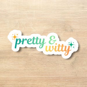 Puede incluir: Una pegatina blanca con las palabras "pretty & witty" en un degradado de verde azulado, verde, amarillo y naranja. La pegatina tiene detalles de estrellas y brillo. La pegatina está sobre una superficie de madera marrón claro.