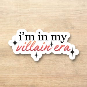Puede incluir: Una pegatina blanca con el texto "i'm in my villain era" en letras negras y degradado rojo-naranja. La pegatina tiene una forma única con cuatro acentos de estrellas negras.