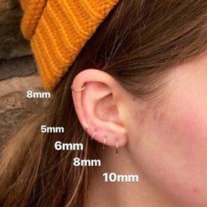 Young woman wearing beanie and leaning against brick wall with multiple upper ear and earlobe piercings filled with seamless hoops in graduating sizes including 5mm, 6mm, 8mm and 10mm.