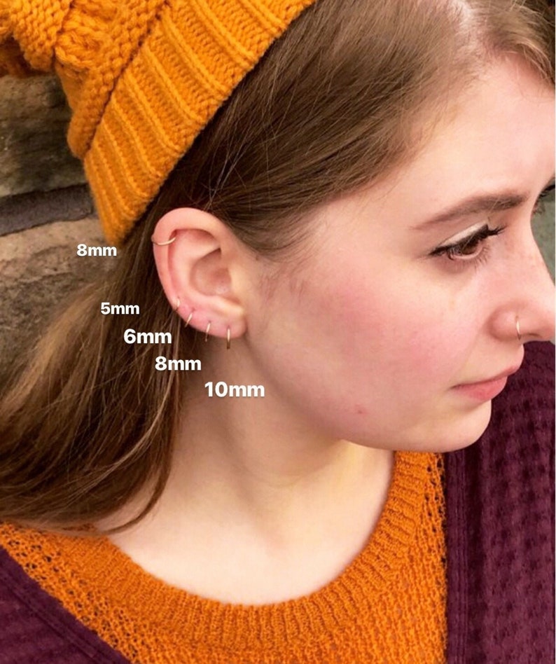 Young woman wearing beanie and leaning against brick wall with multiple upper ear and earlobe piercings filled with seamless hoops in graduating sizes including 5mm, 6mm, 8mm and 10mm.