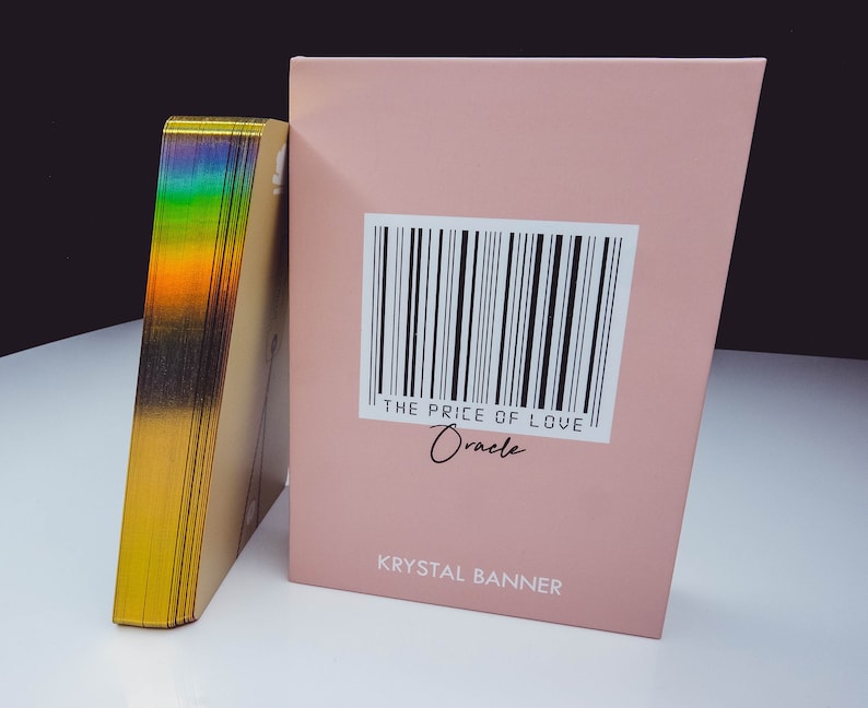 Puede incluir: Una caja rosa con un c&oacute;digo de barras y el texto "THE PRICE OF LOVE Oracle" y "KRYSAL BANNER". La caja est&aacute; al lado de una pila de cartas doradas con un efecto arco&iacute;ris iridiscente.
