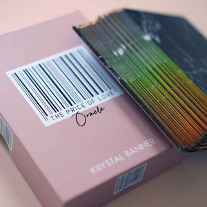 Puede incluir: Una caja rosa con un c&oacute;digo de barras y el texto "THE PRICE OF LOVE Oracle" y "KRYSAL BANNER". La caja est&aacute; abierta y se ve una baraja de cartas con un dise&ntilde;o negro e iridiscente arco&iacute;ris.