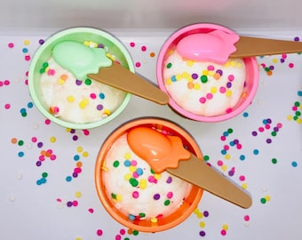 Peut inclure: Trois petits bols de crème glacée blanche avec des pépites colorées, chacun avec une cuillère en plastique et un manche brun. Les bols sont sur une surface blanche avec des pépites colorées éparpillées autour.