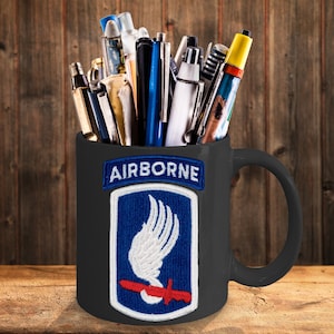 May include: Black ceramic mug with a white and blue airborne insignia and the word "AIRBORNE" in white letters. The mug is filled with pens and pencils.