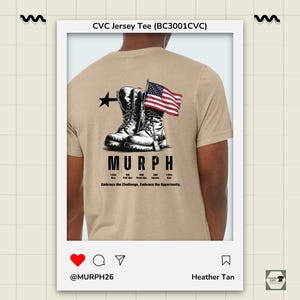 May include: Heather tan CVC jersey tee featuring a graphic of combat boots, an American flag, and the word "MURPH." The design includes a black star and text detailing a workout routine. The shirt also has the phrase "Embrace the Challenge. Embrace the Opportunity."