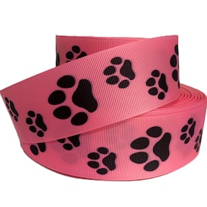 May include: A roll of pink ribbon with a repeating pattern of black paw prints. The ribbon is made of a textured material and is ideal for crafting or gift wrapping. The paw print design adds a playful touch.