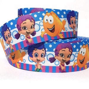 May include: Blue and white ribbon with a repeating pattern of cartoon characters from the animated series Bubble Guppies. The characters include a purple-haired girl, a blue-haired boy, and a yellow fish. The ribbon has a pink and white striped pattern.