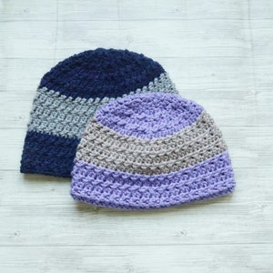 May include: Two crocheted hats, one in a dark blue and gray color combination and the other in a light purple and gray color combination.
