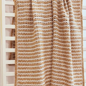 May include: A handmade, rectangular crochet blanket in a warm, neutral colour palette. The blanket features a striped pattern with alternating rows of tan and off-white. The texture is visible, and the blanket is draped over a white cot rail.