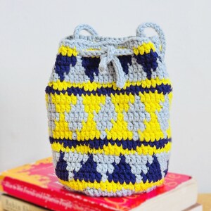 May include: A small crocheted drawstring bag with a geometric pattern in shades of gray, yellow, and blue. The bag is sitting on top of two books.