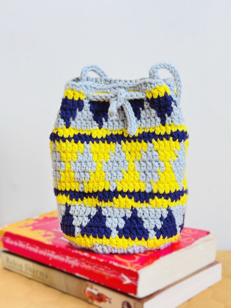 May include: A small crocheted drawstring bag with a geometric pattern in shades of gray, yellow, and blue. The bag is sitting on top of two books.