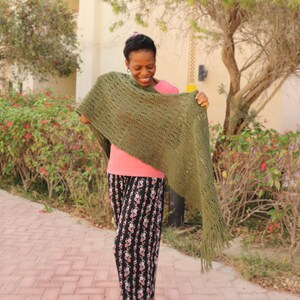 May include: A woman wearing a green crochet shawl with fringe. The shawl is open at the front and has a lacy pattern. The woman is wearing a pink shirt and black and white floral trousers.
