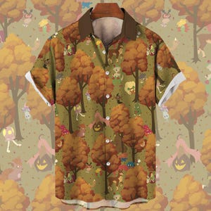 Op de afbeelding: Korte mouwen button-down shirt met een grillige herfstbosprint. Het shirt heeft een bruine kraag en manchetten, met een patroon van cartoonbomen, wezens en pompoenlantaarns op een groene achtergrond. De mouwen hebben witte manchetten.