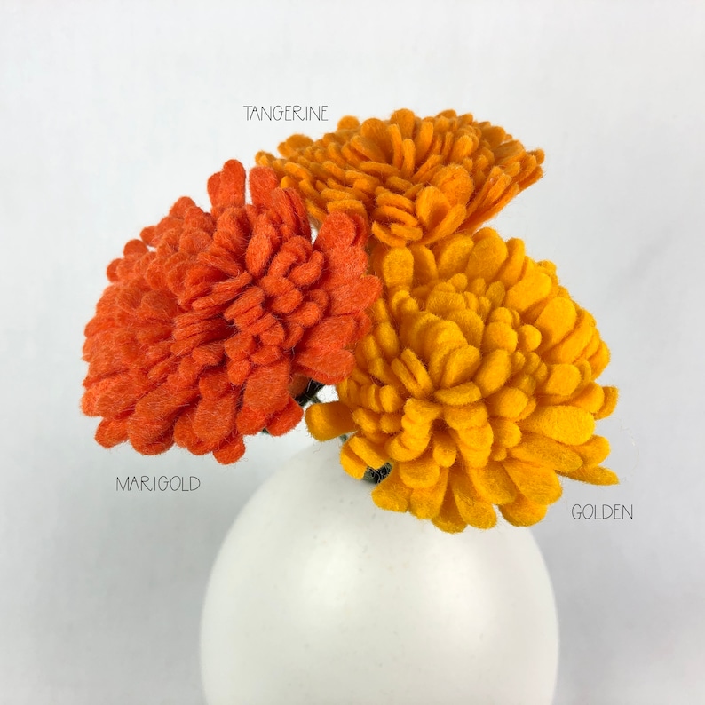 May include: Three felt flower stems in a white vase. The flowers are orange, yellow, and a darker orange. The flowers are labeled "Tangerine", "Marigold", and "Golden".