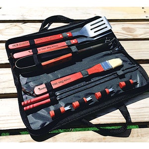 Puede incluir: Un juego de herramientas para barbacoa negro con herramientas con mango rojo, que incluyen una espátula, pinzas, un cuchillo, un cepillo y brochetas. El kit incluye el texto "OUR DADDY", "LIPPIN AMAZING" y "IS THE BBQ KING!"