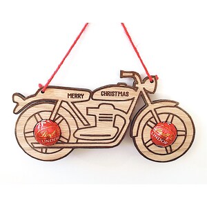 May include: Wooden motorcycle Christmas ornament with the words "MERRY CHRISTMAS" etched on it. The ornament has two Lindor chocolate candies attached to the wheels and a red string for hanging.