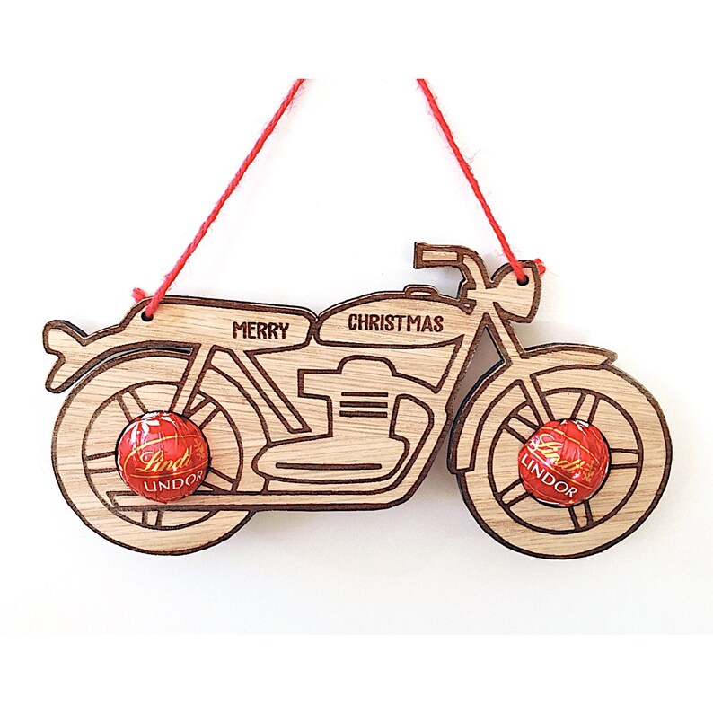 May include: Wooden motorcycle Christmas ornament with the words "MERRY CHRISTMAS" etched on it. The ornament has two Lindor chocolate candies attached to the wheels and a red string for hanging.