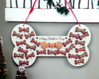 May include: A white, bone-shaped Christmas advent calendar for dogs, with 24 numbered slots holding dog biscuits. The calendar has a red ribbon for hanging and the text "Merry Christmas Tansy". The biscuits are brown with red and green ribbon accents.