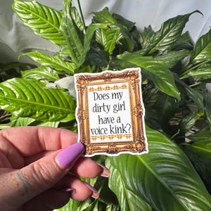 Puede incluir: Una pegatina con un marco dorado ornamentado y el texto "Does my dirty girl have a voice kink?" se sostiene frente a una exuberante planta verde. El marco de la pegatina tiene un diseño floral decorativo.