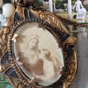May include: Vintage gold-toned oval pendant necklace with a sepia-toned image of two figures. The pendant has ornate detailing and is attached to a delicate chain with white beads. The image is framed by a decorative border.